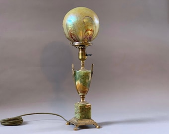 May include: A green and gold table lamp with a round iridescent glass shade. The lamp has a base made of green marble with gold accents.
