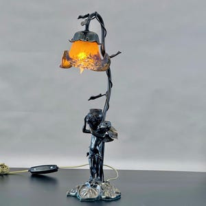 May include: A black metal table lamp with a floral design and a cherub holding a vase. The lampshade is a frosted glass flower with a yellow glow.