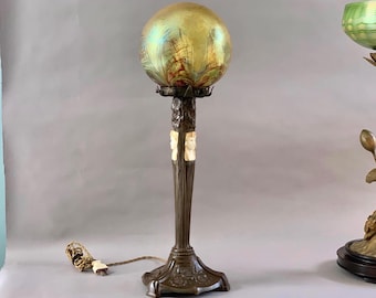 Op de afbeelding: Een bronzen tafellamp met een ronde, iriserende glazen kap. De lamp heeft een decoratieve voet met een gesneden ontwerp en een witte marmeren accent.