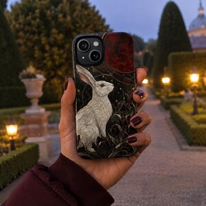 May include: A smartphone case featuring a detailed illustration of a white rabbit sitting among dark, stylized flowers. The background is black with gold star accents and a large, textured red circular element at the top, creating a celestial theme.