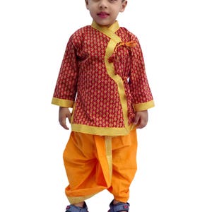 May include: A young boy wearing a red and gold patterned kurta with a yellow dhoti and blue sandals. The kurta has a yellow trim and a tassel.
