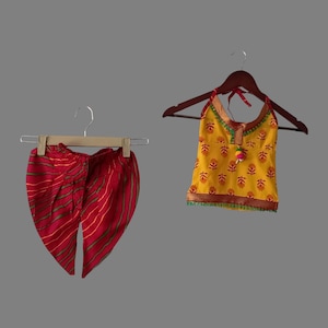 Op de afbeelding: Een tweedelige outfit voor een jong meisje. Het bovenstuk is geel met een bloemenprint en een groene rand. Het onderstuk is rood met groene strepen.