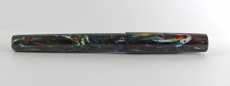 May include: A colorful, marbled fountain pen with a green, blue, red, and white pattern. The pen is closed and lying on a white surface.