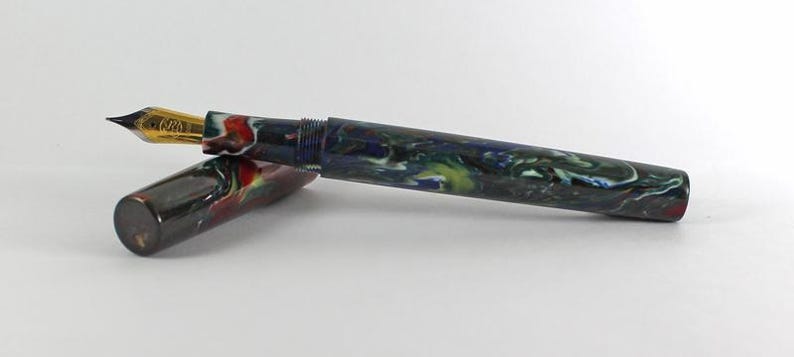 May include: A black and blue fountain pen with a gold nib and a colorful marbled design. The pen is capped and lying on a white surface.