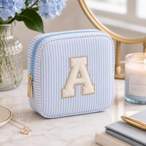 May include: A square, zippered travel jewelry case featuring a light blue and white pinstripe pattern. The front of the case is adorned with a large, white, varsity-style letter A patch with a gold-colored border. The case has a light blue zipper.