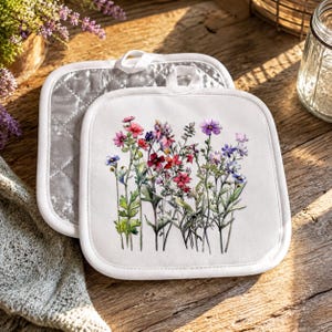 May include: Two square white pot holders featuring a watercolor-style illustration of various wildflowers in shades of pink, red, purple, and blue. The front pot holder displays the floral print, while the back one shows a quilted silver texture.