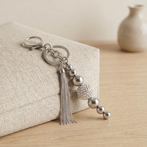 May include: A silver-toned keychain featuring a circular ring, a lobster clasp, and a dangling charm assembly. The charm consists of a fine chain tassel and a vertical arrangement of smooth metallic spheres interspersed with one crystal-encrusted sphere.