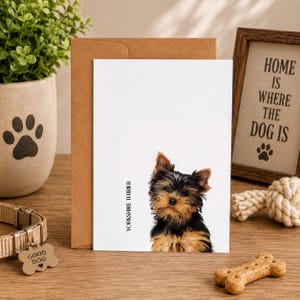 a greeting card with a Yorkshire Terrier dog on it, placed on a wooden surface. The card is accompanied by a brown envelope, a potted plant, a dog toy, and a framed sign with the text "HOME IS WHERE THE DOG IS".