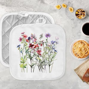 May include: Two white square pot holders. One has a quilted gray back and a white loop. The other has a watercolor floral design with pink, purple, and blue flowers. A cup of coffee, a small pie, and other food items are on the table.