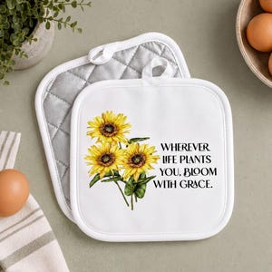 May include: A square white quilted pot holder featuring a printed illustration of three yellow sunflowers with green leaves and the black text that reads: WHEREVER LIFE PLANTS YOU, BLOOM WITH GRACE. The pot holder has a white hanging loop at the top.