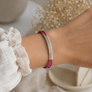 May include: A close-up of a bracelet with three silver-colored metal tubes and a magenta-colored band. The bracelet is worn on a wrist. The background is blurred, with a white top and dried flowers.