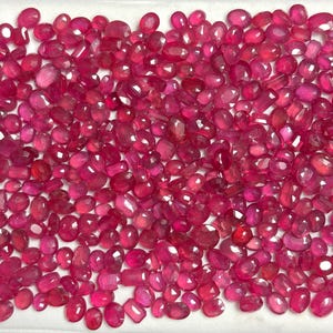 May include: A collection of faceted rubies in shades of red and pink, with oval and rectangular shapes. The gemstones are scattered on a white surface, displaying their polished surfaces and clarity. The arrangement highlights the variety of shapes and sizes of the gemstones.