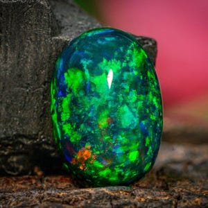 May include: An oval opal gemstone displaying a vibrant array of colours. The gem features a mix of green, blue, and orange hues, set against a dark background. The opal is displayed on a textured, dark surface.