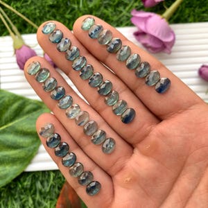 May include: Several faceted, oval gemstones in varying shades of blue and green are displayed. The gems are held in a hand, showing their clarity and cut. The stones are arranged in rows, highlighting their individual facets and overall brilliance. The background is blurred, focusing on the gemstones.