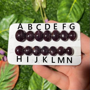 May include: A white card with the letters A through G and H through N printed on it. There are 14 dark red round gemstones arranged in two rows on the card.