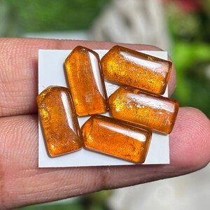 May include: A collection of four polished, rectangular amber gemstones with rounded edges. Each piece displays a translucent, deep orange hue with visible internal inclusions and a smooth, reflective surface, arranged on a small white square.