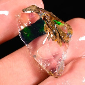 Könnte beinhalten: Ein roher, durchscheinender Opal-Edelstein mit einem dunkelgrünen und blauen Innenmuster. Der Opal hat eine raue, braune Matrix an einer Kante und zeigt schillernde Farbtöne.