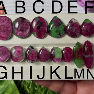 May include: A collection of ruby gemstones in various shapes, including oval, teardrop, and elongated forms. The gemstones display a mix of deep red and green hues, with visible inclusions. The gemstones are arranged on a white display board, labeled with black capital letters.