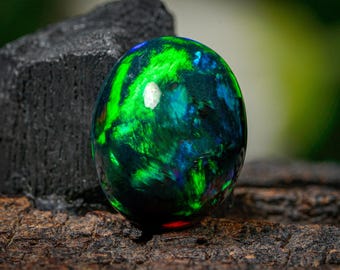 May include: A polished, oval-shaped gemstone with a vibrant display of color. The stone features swirling patterns of green, blue, and black, with a bright, reflective surface. The gemstone is set against a dark, textured background.