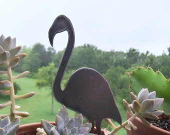 May include: A metal flamingo garden stake stands in a terracotta pot filled with succulents. The flamingo is dark gray and has a long, curved neck. The succulents are light green and gray, with water droplets on the leaves. The background is a blurred view of green trees.
