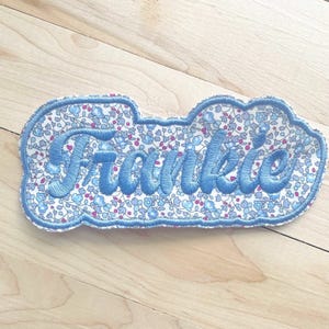 May include: A cloud-shaped embroidered patch with the name "Frankie" in light blue script. The patch has a floral pattern in blue and pink on a white background, with a blue stitched border.