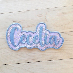 May include: A custom name patch featuring the name Cecelia embroidered in blue thread. The lettering is set against a light purple background with a white floral pattern, finished with a blue stitched border that follows the shape of the text.