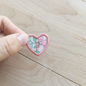 May include: A heart-shaped fabric patch with a floral pattern in pink, green, and white. The patch has a pink embroidered border and the letter "P" in the center. The patch is held by a hand.