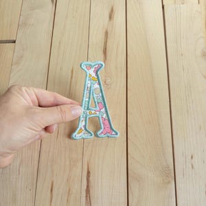 Peut inclure: Un écusson brodé en forme de lettre A avec un motif floral composé de fleurs roses, jaunes et bleues sur fond blanc, bordé d'une couture bleu clair. L'écusson est présenté sur une surface en bois de couleur claire.
