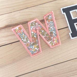 May include: A fabric letter W patch featuring a floral pattern with shades of pink, yellow, blue, and green, finished with a coral-colored stitched border. A portion of a black letter patch with a white stitched border is visible to the right.