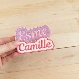 May include: Two embroidered name patches, one lavender with "Esme" in pink script, and one white with "Camille" in pink script. Both patches have a cloud-like shape with a pink border. The patches are held against a light wood background.