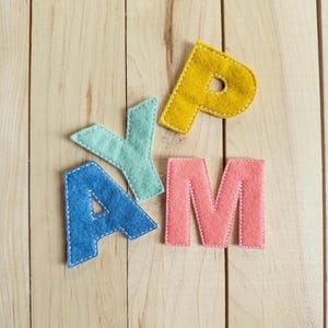 May include: Four individual felt letters spelling the word PLAY, featuring a mustard yellow P, a mint green L, a blue A, and a coral pink Y. Each letter is crafted from soft felt material with visible white stitched borders along the edges.