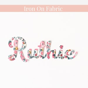 May include: Iron-on fabric letters spelling out the name "Ruthie" in a cursive font. The letters are made of white fabric with a pink and blue floral pattern.
