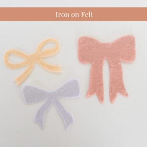 May include: Three iron-on felt bows in different colours: light yellow, light pink, and light purple. The bows are all in the shape of a traditional bow with two loops and a centre knot.
