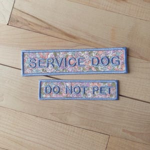 Può includere: Due toppe rettangolari ricamate con un bordo azzurro e un motivo floreale nei toni del rosa, blu e verde. La toppa più grande presenta la scritta SERVICE DOG in blu, mentre la più piccola presenta la scritta DO NOT PET in blu.