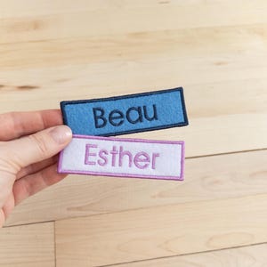 May include: Two embroidered name patches. One is light blue with the name "Beau" in black. The other is white with the name "Esther" in pink. Both patches have a rectangular shape with a colored border.
