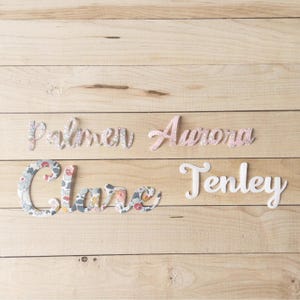 May include: Four wooden name cutouts on a light wood surface. The names are Palmer, Aurora, Clare, and Tenley. Palmer and Clare are covered in a floral fabric. Aurora is a light pink colour, and Tenley is white.