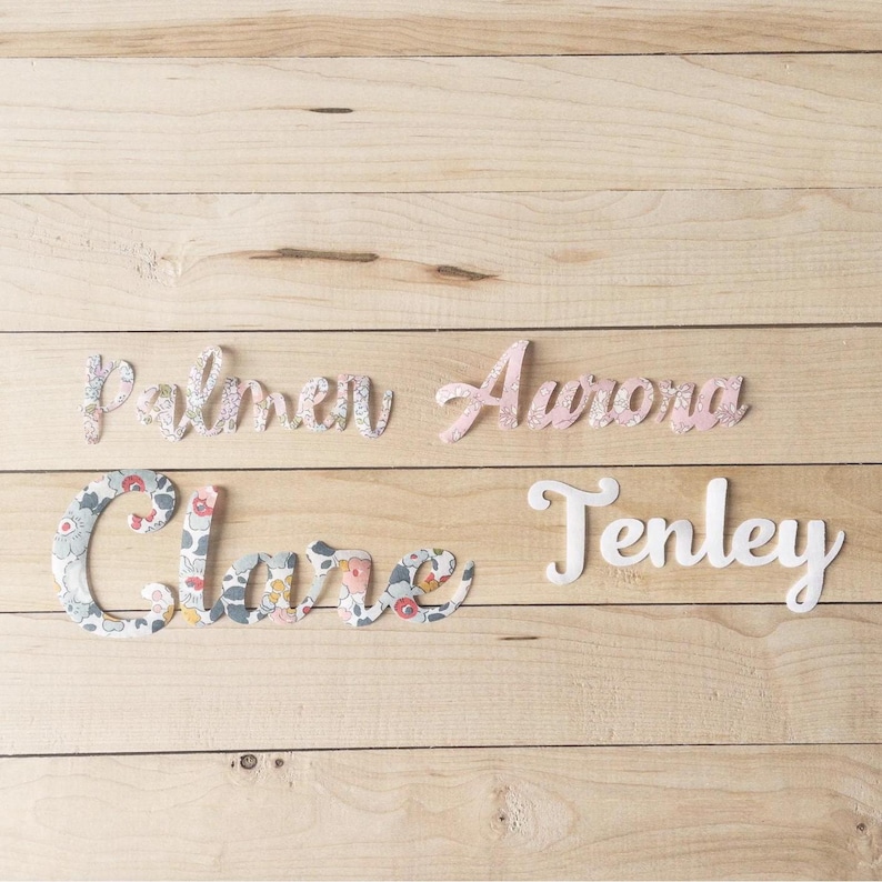 May include: Four wooden name cutouts on a light wood surface. The names are Palmer, Aurora, Clare, and Tenley. Palmer and Clare are covered in a floral fabric. Aurora is a light pink colour, and Tenley is white.