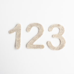 May include: The numbers 1, 2, and 3 are made of a fuzzy, beige material. The numbers are arranged horizontally on a white background.