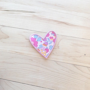 May include: A heart-shaped fabric patch featuring a light pink background with a repeating floral pattern in shades of magenta, light blue, and orange. The patch has a stitched border in a matching light pink thread, creating a clean edge.