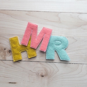 May include: Felt letters spelling "HMR" in yellow, pink, and blue, with white stitching. The letters are arranged on a light wood-grain surface.