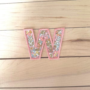 May include: A fabric letter W patch featuring a floral pattern with shades of pink, blue, yellow, and green. The letter is outlined with a thick, stitched pink border, creating a defined edge against the background of the fabric letter.