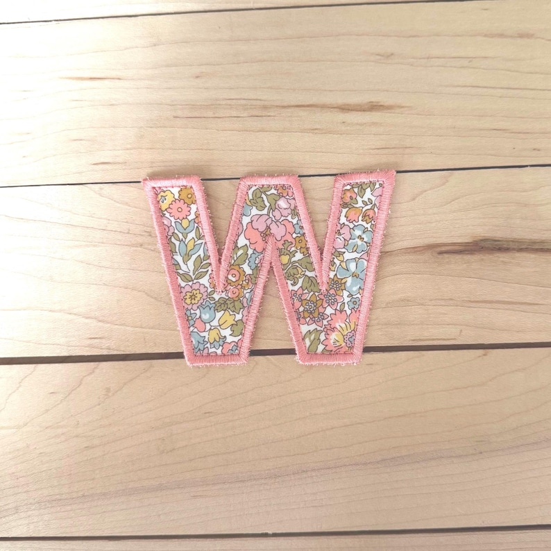 May include: A fabric letter W patch featuring a floral pattern with shades of pink, blue, yellow, and green. The letter is outlined with a thick, stitched pink border, creating a defined edge against the background of the fabric letter.