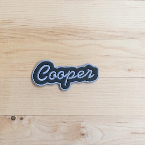 May include: A dark navy blue embroidered patch featuring the name Cooper in a light blue cursive script font. The patch has a distinct, irregular border that follows the shape of the lettering, creating a clean and defined edge for application.