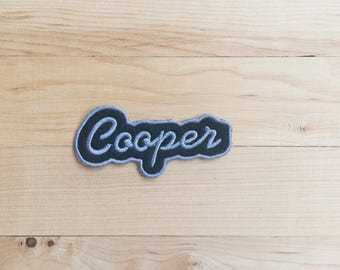 以下が含まれることがあります： 濃いネイビーブルーの刺繍ワッペンで、ライトブルーの筆記体で「Cooper」という名前が記されています。ワッペンは文字の形に沿った独特の不規則な縁取りが施されており、すっきりと明確な輪郭に仕上がっています。