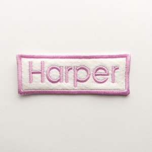 May include: A rectangular embroidered patch featuring the name Harper in pink thread on a white felt background. The patch is bordered by a thick pink stitched edge, creating a clean and defined frame around the centered pink lettering.