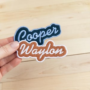 Peut inclure: Deux écussons brodés avec les noms "Cooper" et "Waylon" en écriture cursive. L'écusson "Cooper" est bleu marine avec des coutures bleu clair, tandis que l'écusson "Waylon" est marron avec des coutures blanches. Les deux écussons ont une bordure blanche.
