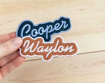 以下が含まれることがあります： スカラップ状の縁取りが施された2枚の刺繍ネームワッペン。上のワッペンはダークネイビー地にライトブルーの筆記体でCooperと刺繍されています。下のワッペンはラストオレンジ地にホワイトの筆記体でWaylonと刺繍されています。どちらも縁がステッチで仕上げられています。