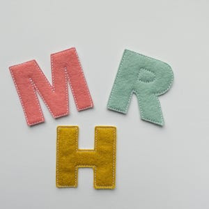 Pode incluir: Três letras de feltro: M em coral, R em verde menta e H em amarelo mostarda. Cada letra é costurada com linha branca nas bordas. As letras estão dispostas sobre um fundo branco.