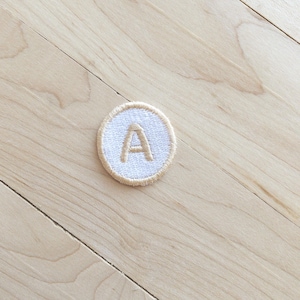 May include: A circular patch with a white centre and a tan border. The letter "A" is embroidered in tan thread in the centre of the patch. The patch is likely an iron-on or sew-on applique for clothing or accessories.