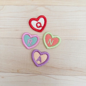 May include: Four embroidered heart-shaped patches in red, pink, green, and purple. Each heart features a different letter: Q, R, N, and A. The patches are on a light wood surface.